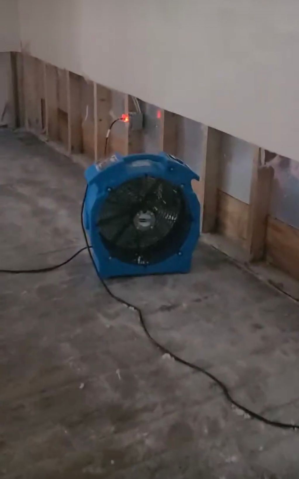 Blue industrial fan plugged in beside an unfinished wall with exposed wooden studs and partially removed drywall.