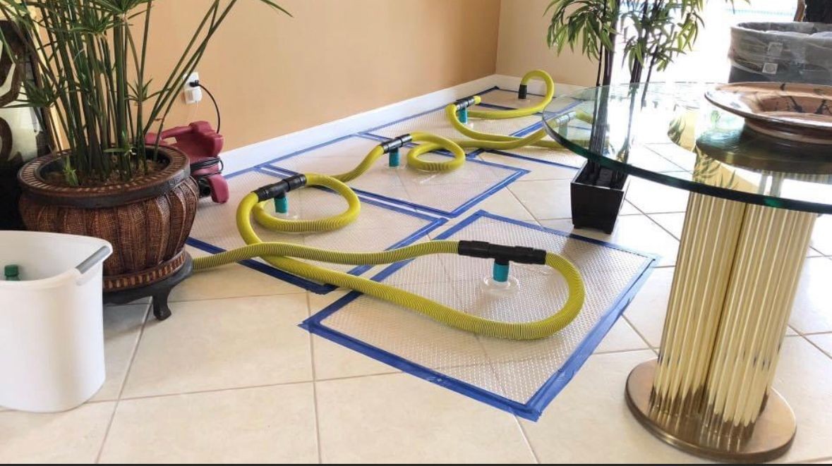 Living room floor with yellow hoses on taped mats for water damage restoration.